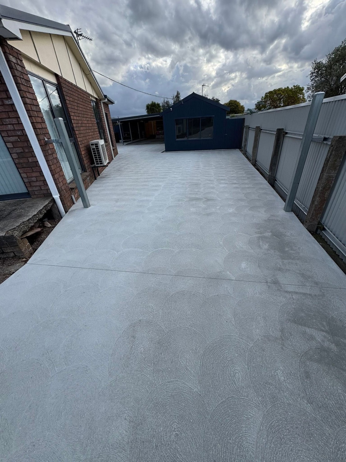 A broom-finished concrete area beside a brick home and navy shed